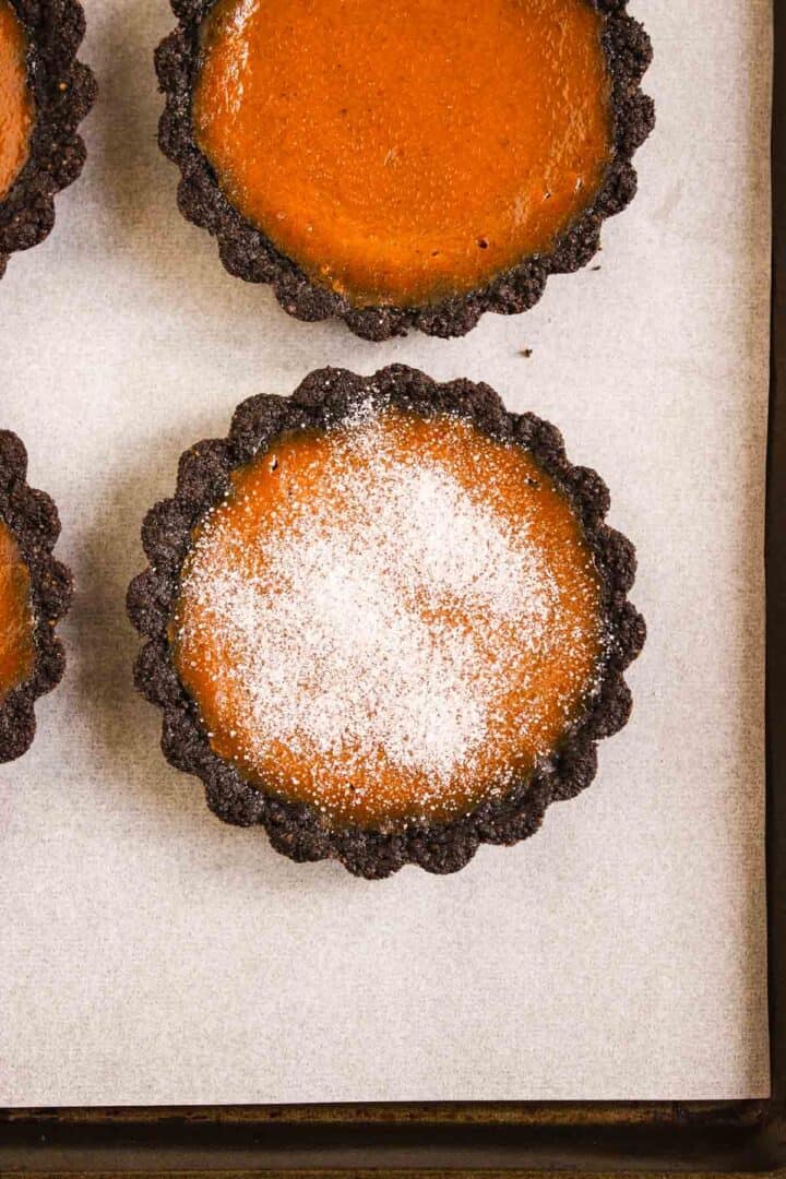 Pumpkin Br&ucirc;l&eacute;e Tarts feature chocolate tart shells filled with rich pumpkin filling, one topped with powdered sugar, all arranged beautifully on parchment paper.