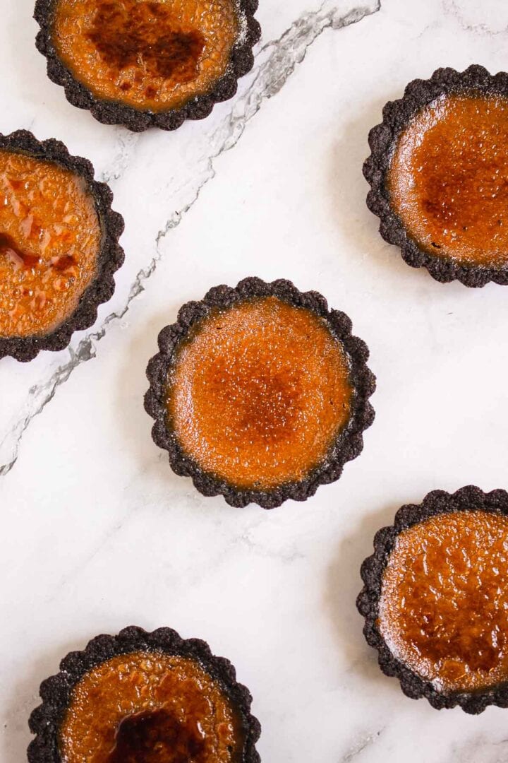 Mini Pumpkin Br&ucirc;l&eacute;e Tarts with caramelized tops and dark crusts sit elegantly on a white marble surface.