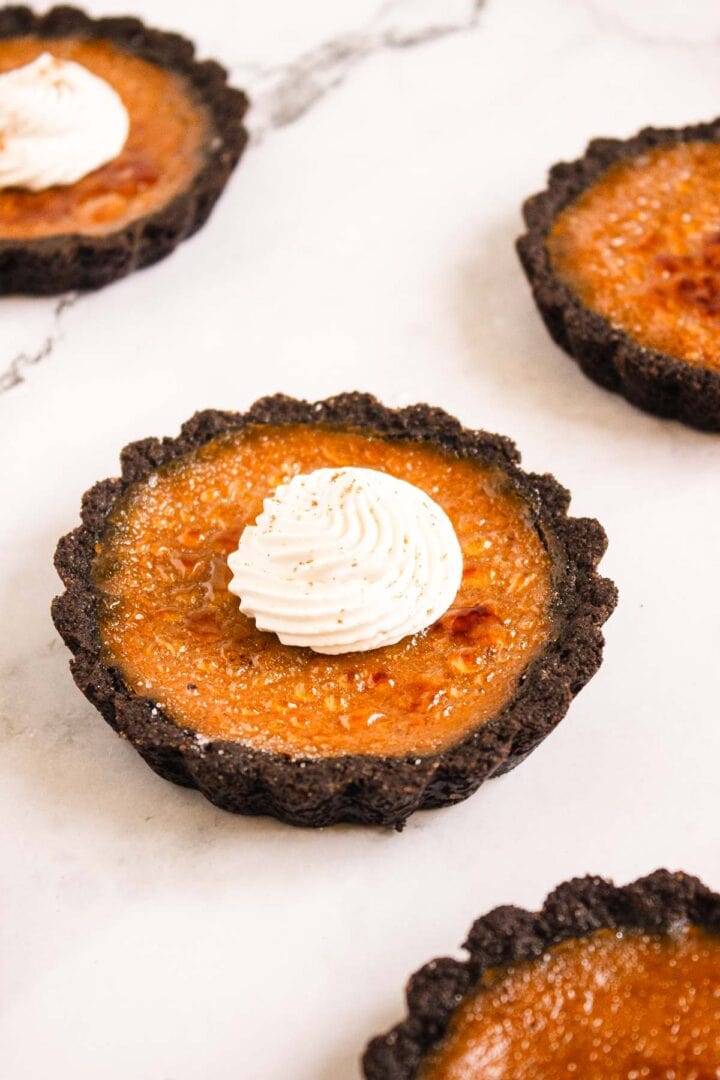 Chocolate tart with caramelized top and a swirl of whipped cream, reminiscent of elegant Pumpkin Br&ucirc;l&eacute;e Tarts, on a light marble surface.