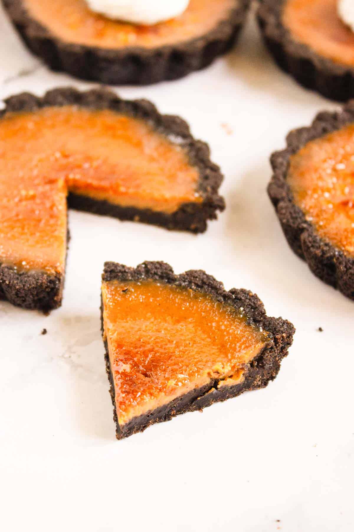 A small tart with a chocolate crust and orange filling, with one slice cut and placed in front&mdash;perfect for pairing alongside Pumpkin Br&ucirc;l&eacute;e Tarts at your dessert table.