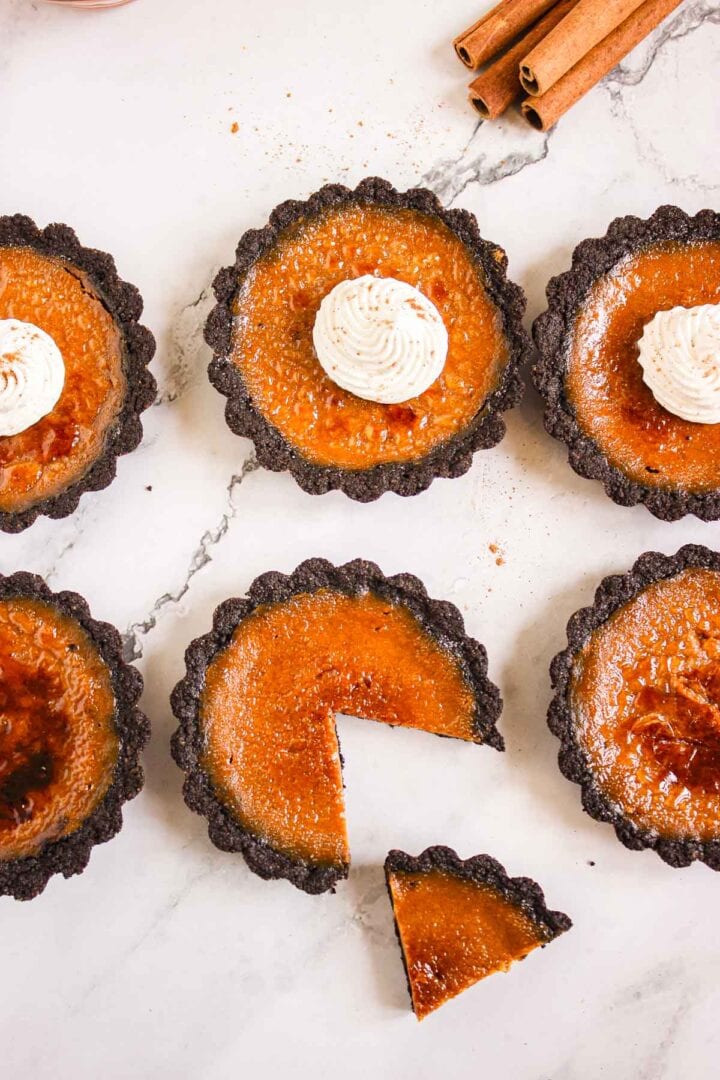 Six Pumpkin Brûlée Tarts with chocolate crusts, one with a slice cut out, each topped with a swirl of whipped cream.