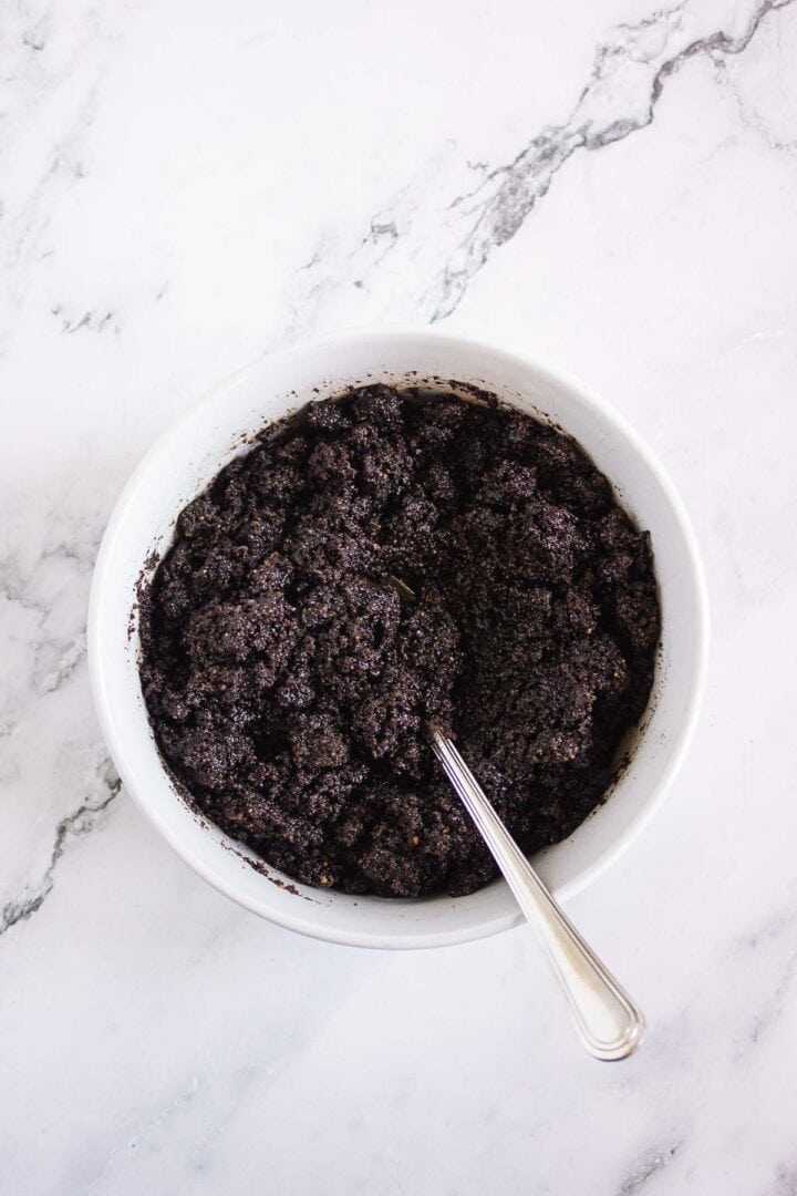 A white bowl with a spoon, filled with a dark, crumbly mixture on a marble surface&mdash;perfect for making Pumpkin Br&ucirc;l&eacute;e Tarts.