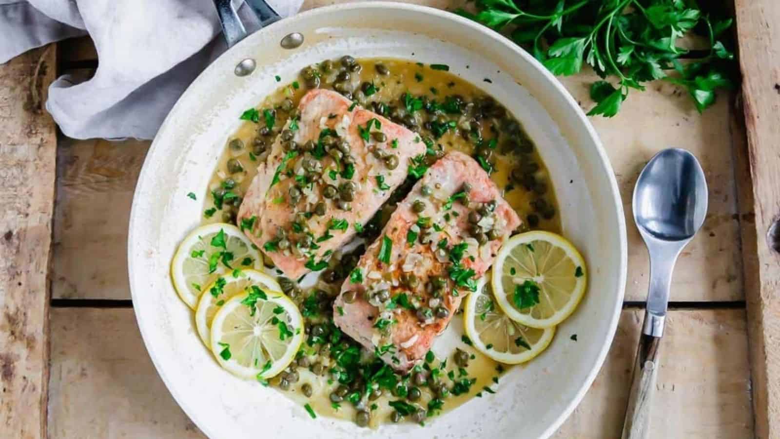 Two cooked salmon fillets in a white skillet with caper sauce, garnished with lemon slices and chopped parsley, next to a metal spoon and fresh parsley on a wooden surface.