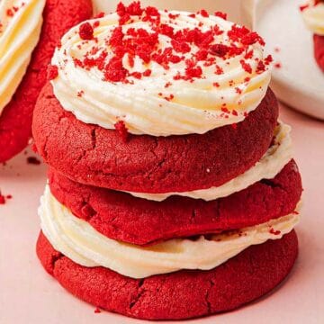 Three stacked red velvet cookies with white frosting and red crumbs, on a pink surface.