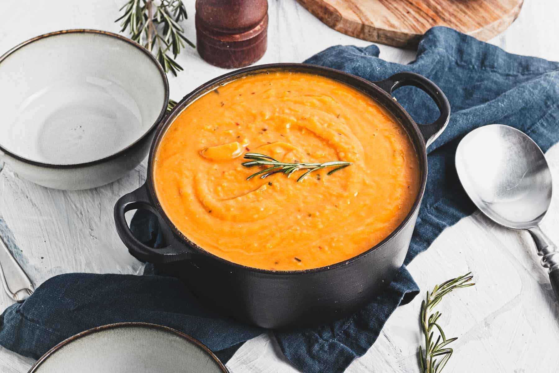 Creamy orange soup in a black pot, garnished with rosemary, beside empty bowls and a spoon on a blue cloth.