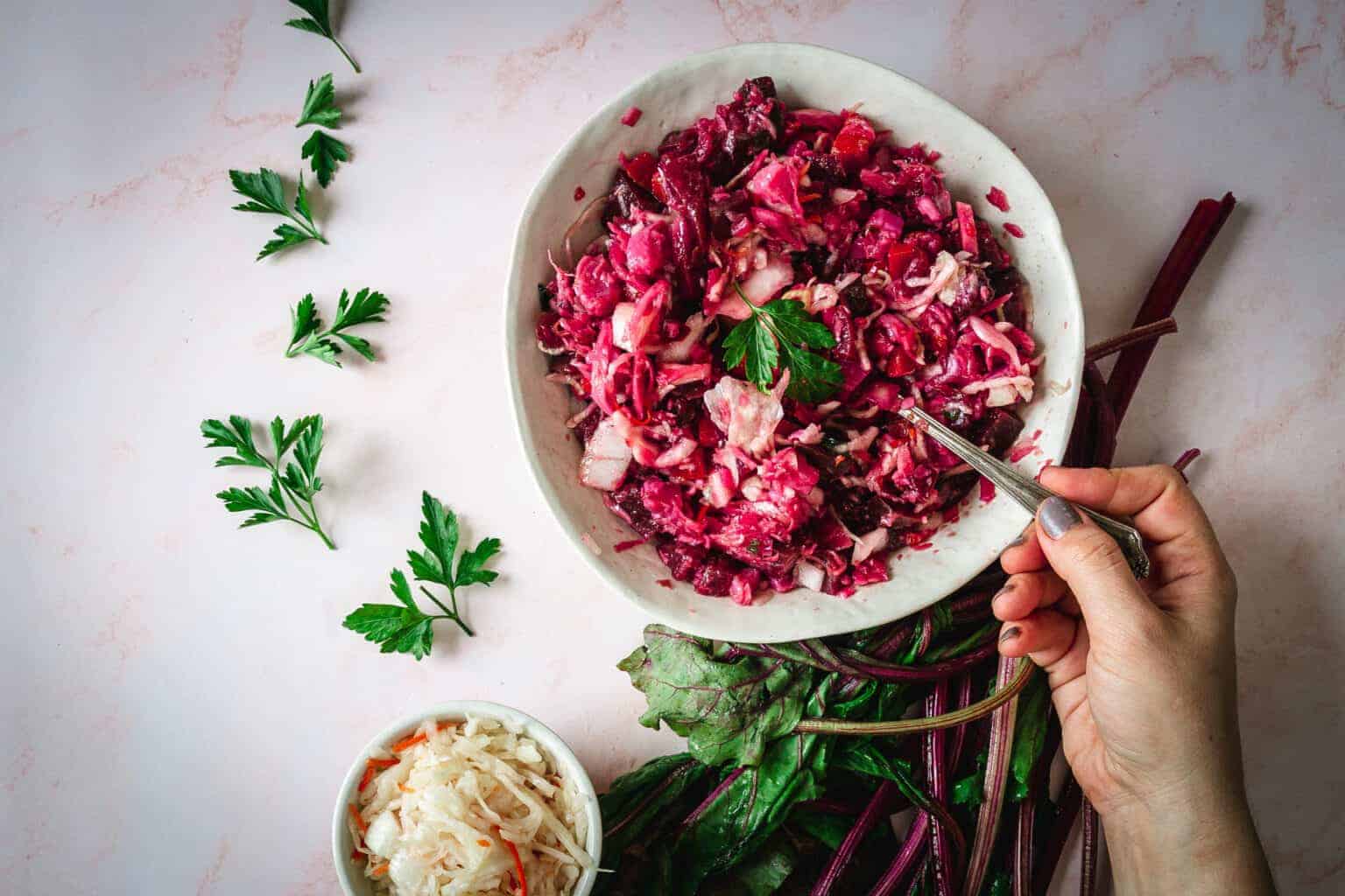 A hand holds a fork in a bowl of beet and cabbage salad, with greens and parsley on a light table.
