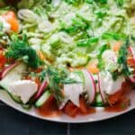 A vibrant Salmon Wreath featuring smoked salmon, cucumber, radish, capers, cream cheese, and fresh dill, served with a side of guacamole dip.