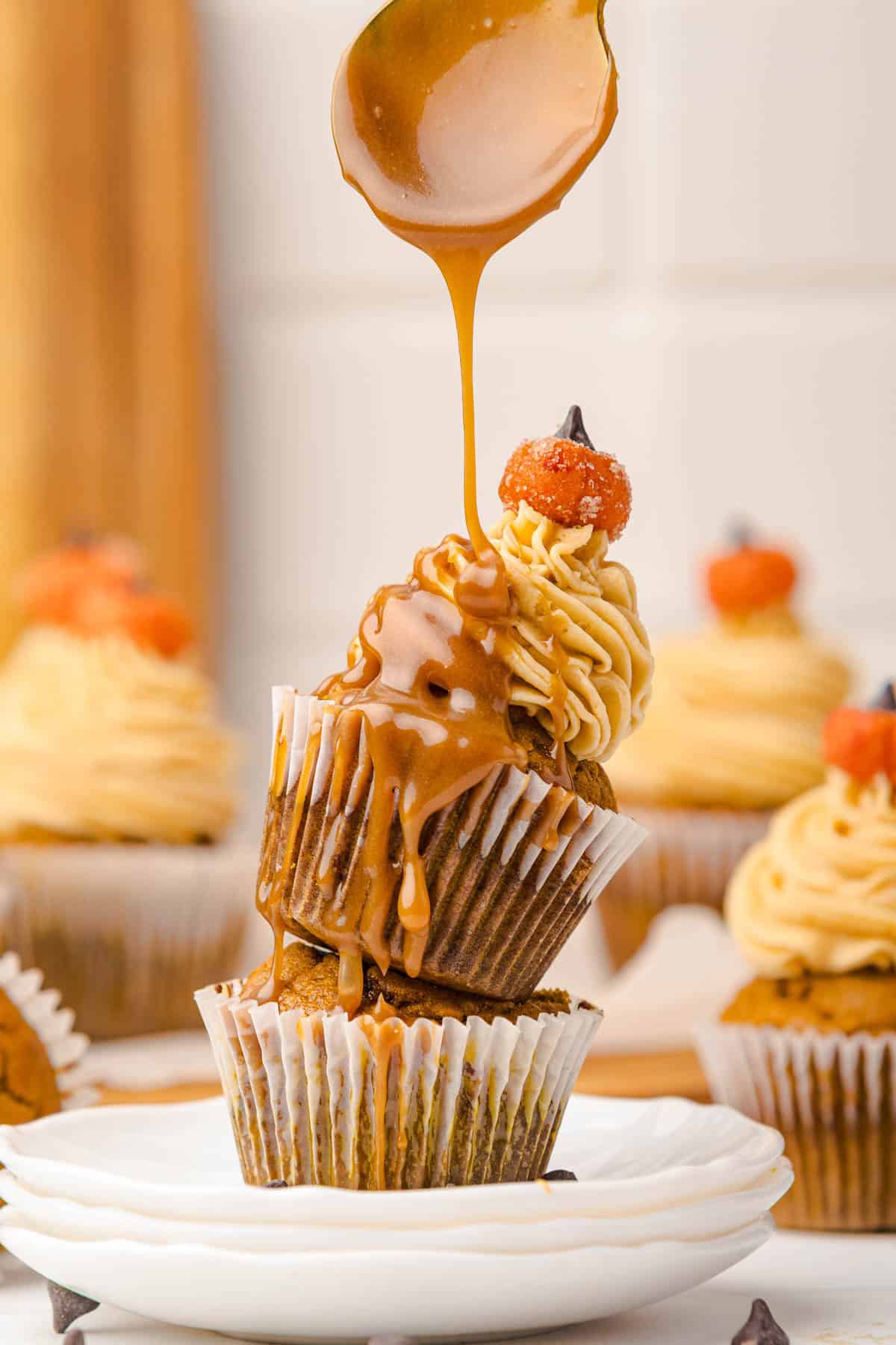 Caramel sauce being poured over stacked Salted Caramel Pumpkin Muffins topped with swirled frosting and pumpkin-shaped decorations.