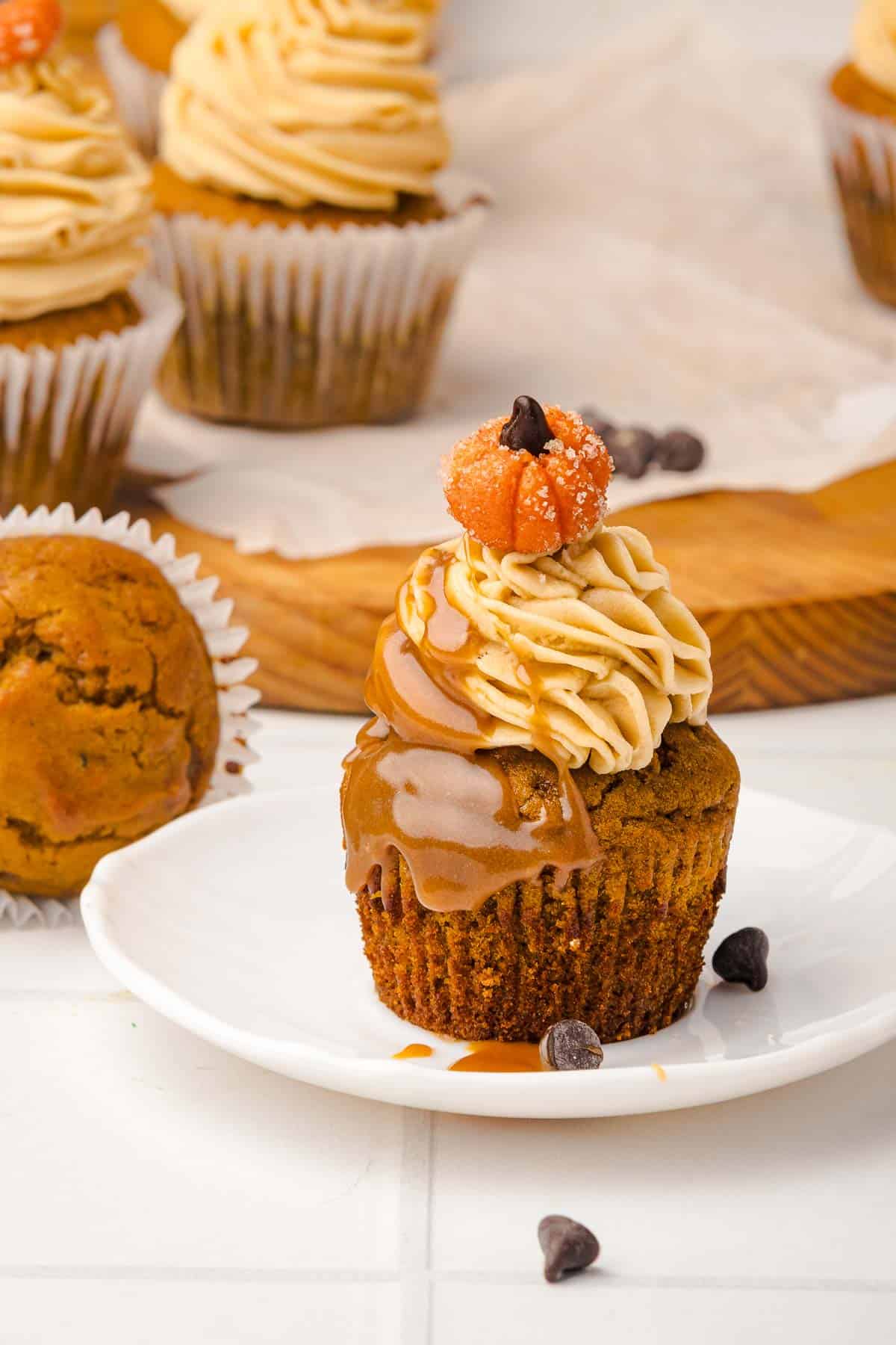 Pumpkin cupcake with caramel drizzle, frosting, a small pumpkin candy, and chocolate chips on a white plate&mdash;just like delightful Salted Caramel Pumpkin Muffins in cupcake form.