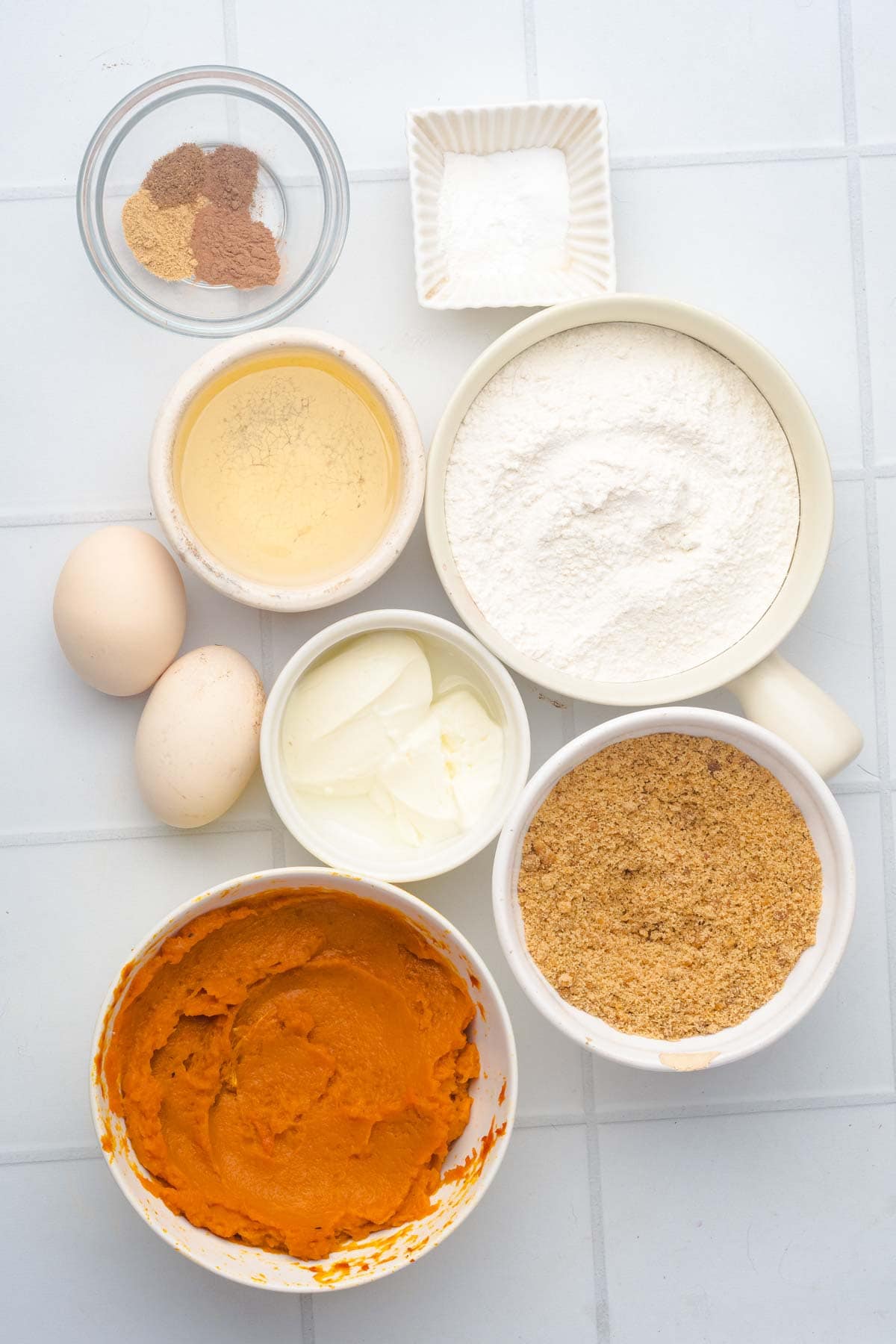 Bowls of flour, sugar, spices, baking powder, eggs, yogurt, and pumpkin puree on a white tiled surface&mdash;perfect for making Salted Caramel Pumpkin Muffins.