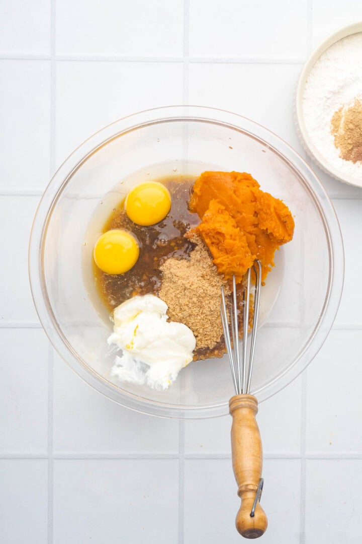 A bowl with eggs, pumpkin puree, yogurt, and sugar&mdash;ingredients for delicious Salted Caramel Pumpkin Muffins&mdash;sits next to a whisk and a bowl of flour and spices.