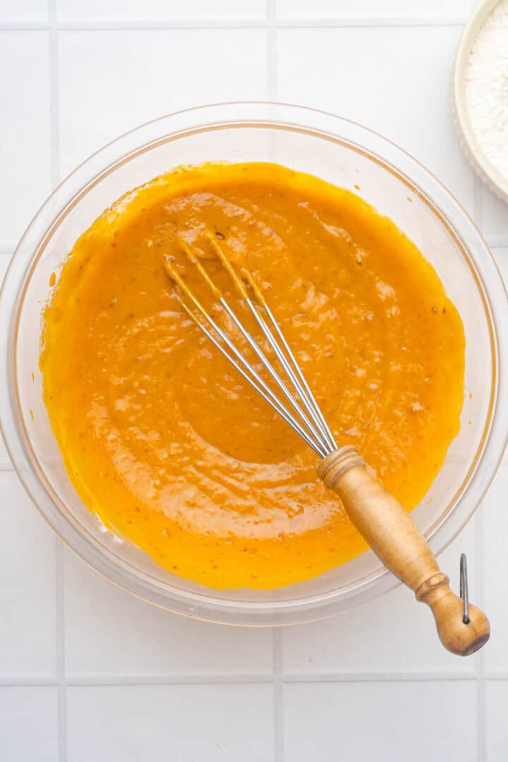 A glass bowl of orange batter for Salted Caramel Pumpkin Muffins sits with a whisk on a white tiled surface.