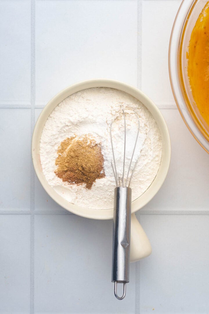 White bowl with flour, spices, and a whisk on a white tiled surface, next to a bowl of orange batter for Salted Caramel Pumpkin Muffins.