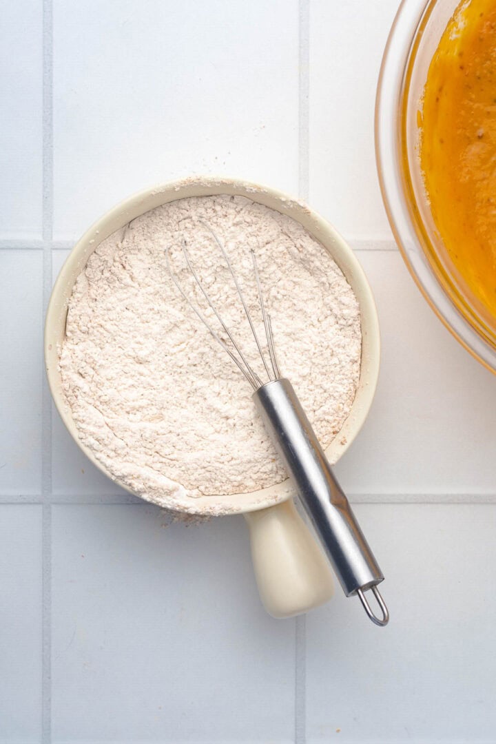 A whisk rests on a bowl filled with flour next to a bowl of orange batter, ready for baking delicious Salted Caramel Pumpkin Muffins on a white tiled surface.
