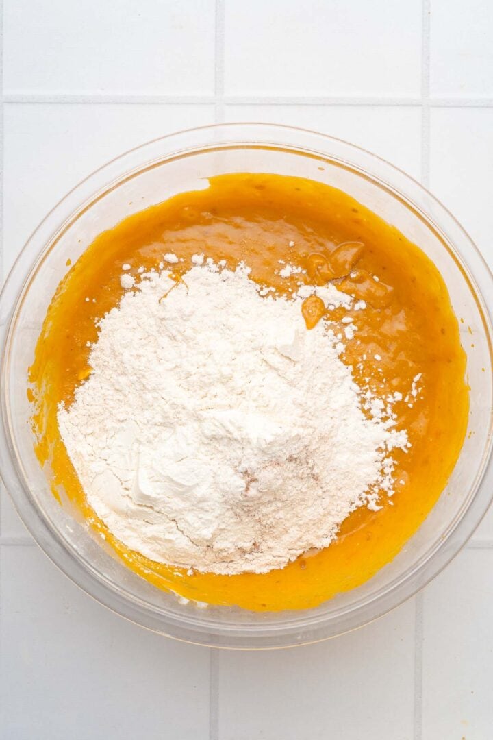 A glass bowl with flour on top of an orange batter for Salted Caramel Pumpkin Muffins, sitting on a white tiled surface.