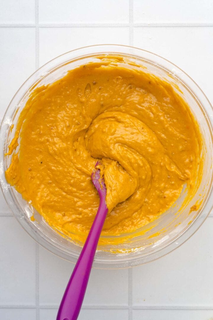 A glass bowl of pumpkin batter for Salted Caramel Pumpkin Muffins is being mixed with a purple spatula on a white tiled surface.