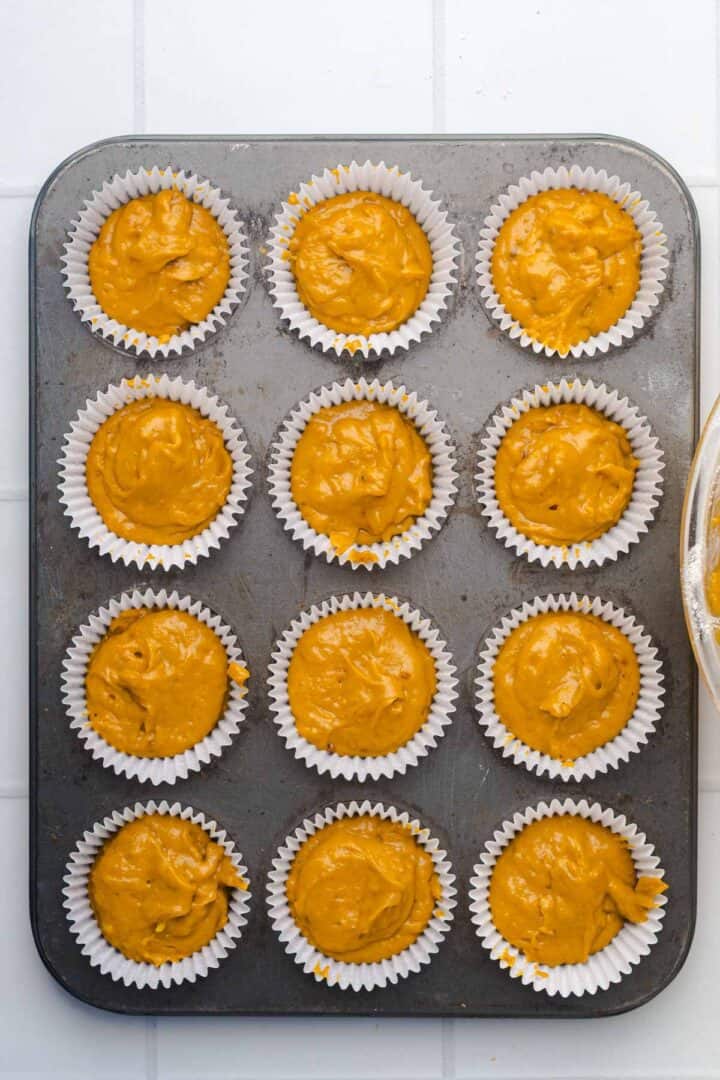 A muffin tin filled with paper liners, each containing Salted Caramel Pumpkin Muffins batter, ready to be baked.