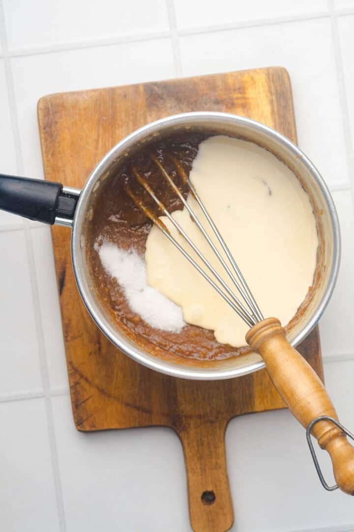 A whisk in a saucepan mixing brown and white batter for Salted Caramel Pumpkin Muffins, on a wooden board atop a white tiled surface.