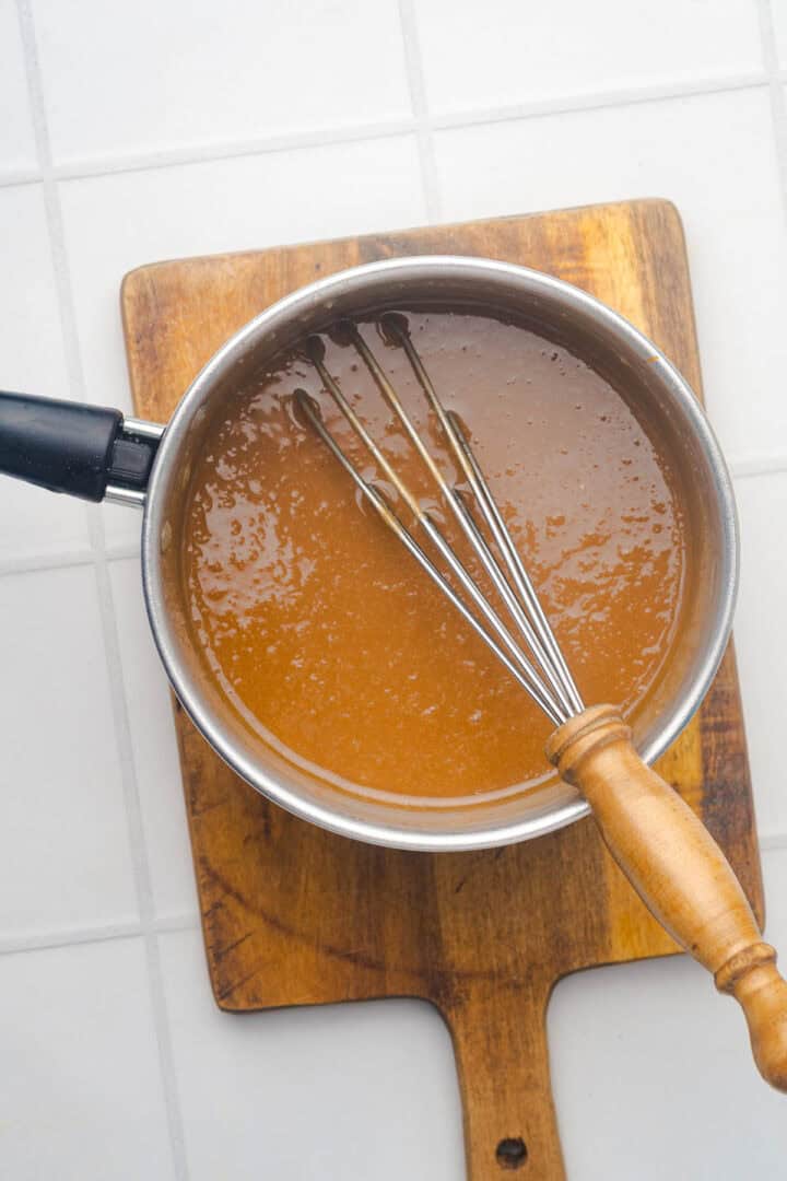A saucepan of brown gravy with a whisk rests on a wooden board, set atop a white tiled surface&mdash;perfect for pairing with salted caramel pumpkin muffins.