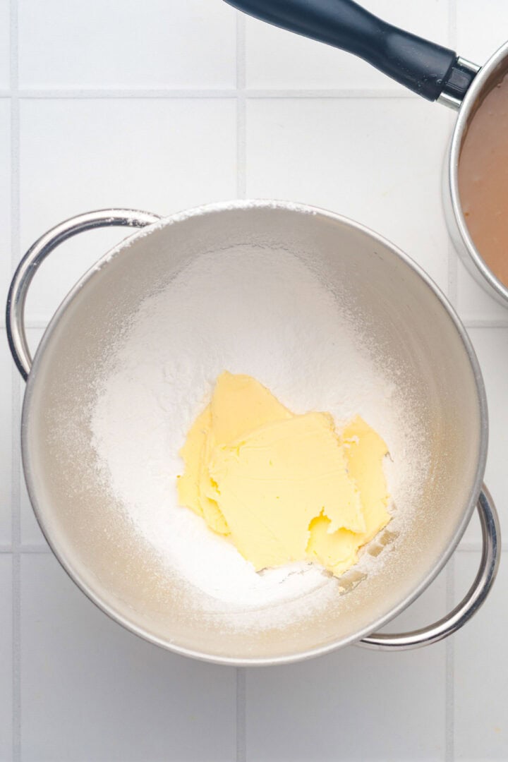 A metal bowl with flour and butter sits on a white tiled surface, next to a pan with a dark mixture&mdash;ingredients ready for baking Salted Caramel Pumpkin Muffins.