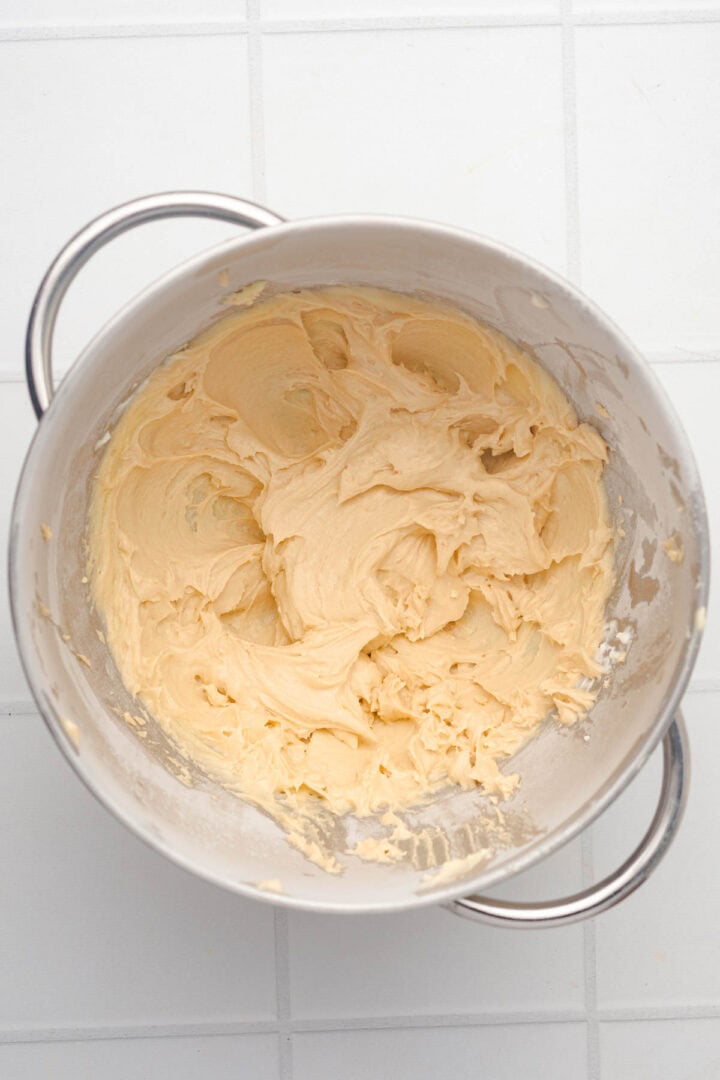 A mixing bowl with creamy, light yellow cake batter for Salted Caramel Pumpkin Muffins sits on a white tiled surface.