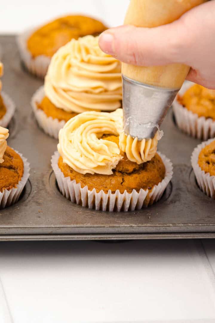 A hand piping frosting onto a Salted Caramel Pumpkin Muffin in a muffin tray with other frosted cupcakes.