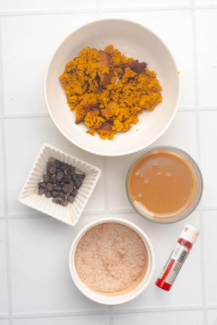 Four bowls with various ingredients for Salted Caramel Pumpkin Muffins and a small tube of food coloring sit on a white tiled surface.