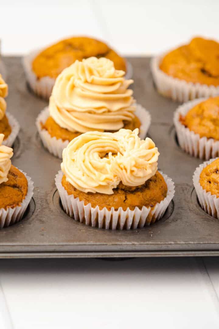 Salted Caramel Pumpkin Muffins with swirled frosting sit in a metal muffin tin on a white surface.