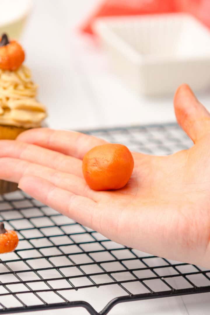A hand holding a small orange ball of fondant over a cooling rack, with Salted Caramel Pumpkin Muffins and a cupcake in the background.