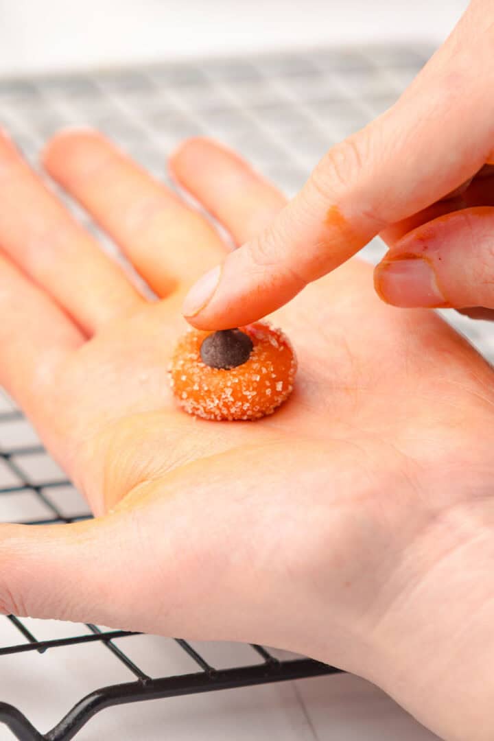 A hand pressing a chocolate chip into a sugar-coated cookie dough ball on a cooling rack, evoking the warm, cozy feeling of baking Salted Caramel Pumpkin Muffins in the fall.