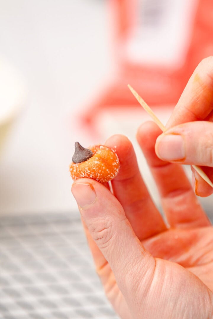 A hand holds a small pumpkin-shaped candy with a chocolate chip stem, using a toothpick for detail&mdash;perfect alongside your favorite Salted Caramel Pumpkin Muffins.