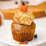 Pumpkin cupcake topped with caramel, piped frosting, and a small candy pumpkin, reminiscent of our Salted Caramel Pumpkin Muffins, served on a white plate.