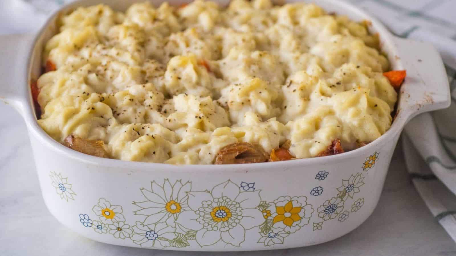 A casserole dish filled with baked shepherd's pie, topped with mashed potatoes and sprinkled with black pepper. The dish has a floral pattern on the side.