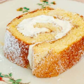 A slice of Swiss roll cake with cream filling, dusted with powdered sugar, on a floral-patterned plate.