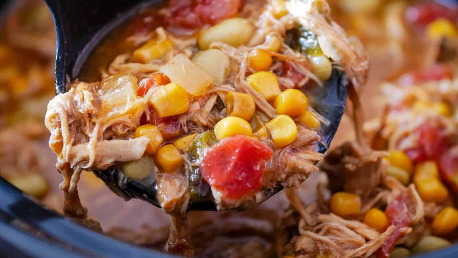 A ladle scooping vegetable stew with corn, tomatoes, beans, and shredded meat from a pot.