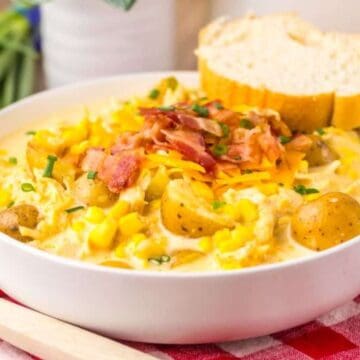 A bowl of creamy potato and corn chowder topped with bacon and cheese, served with bread on a red checkered cloth.