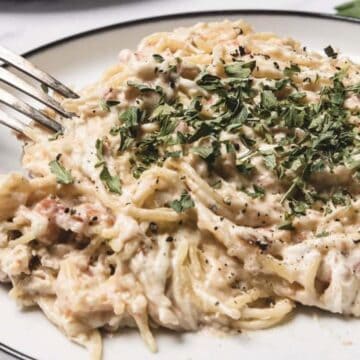 A plate of creamy spaghetti with shredded chicken, topped with chopped parsley and black pepper.