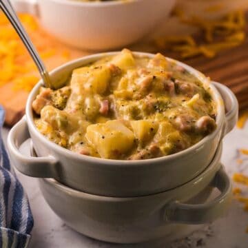 Bowl of cheesy potato soup with a spoon, topped with herbs, beside a striped napkin and shredded cheese.