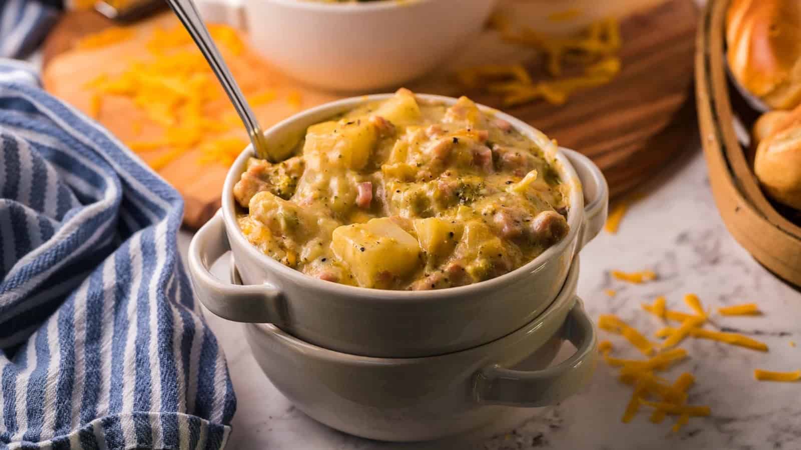 Bowl of cheesy potato soup with a spoon, topped with herbs, beside a striped napkin and shredded cheese.