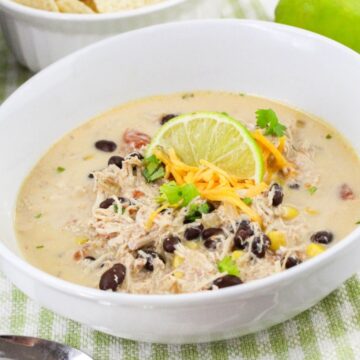 A bowl of creamy chicken soup with black beans, corn, cheese, cilantro, and a lime slice, served with tortilla chips.