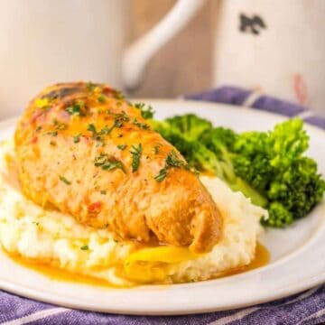 A plate of chicken breast on mashed potatoes with broccoli, garnished with herbs.