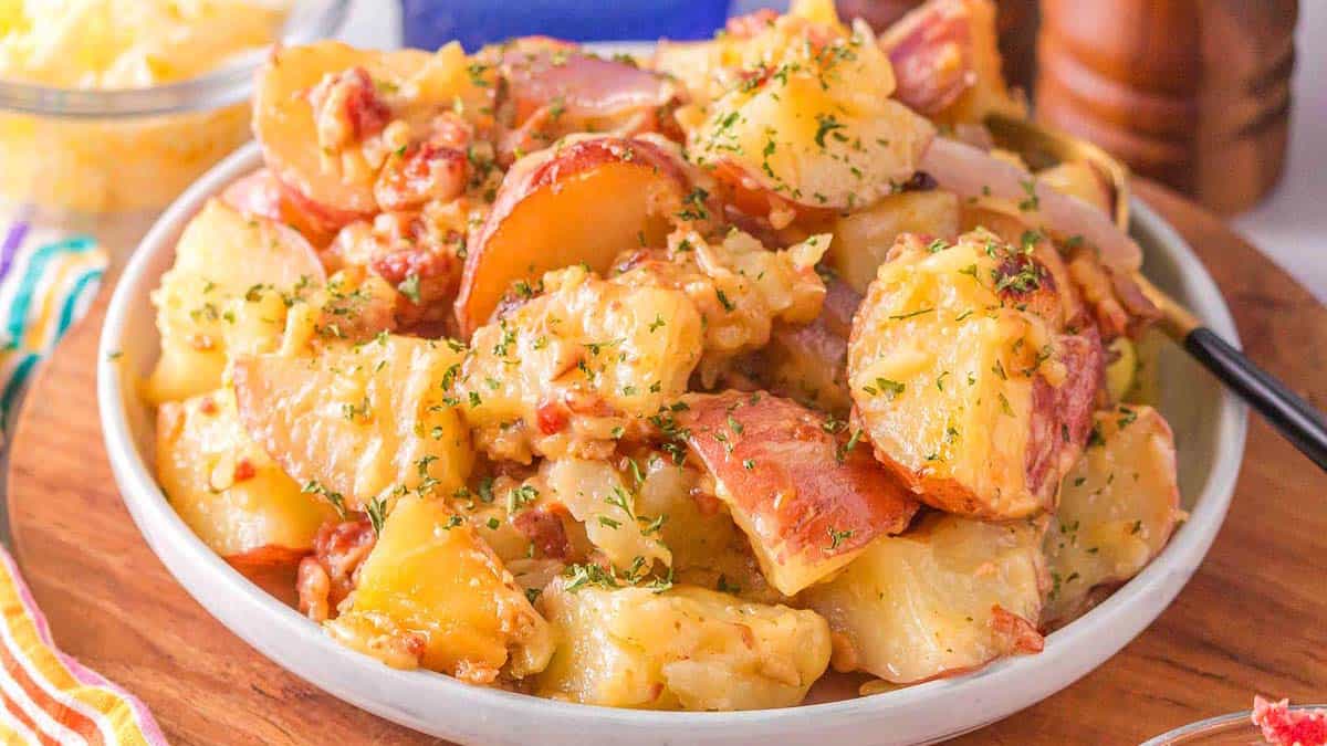 A bowl of loaded potato salad with cheese, bacon, and herbs on a wooden table.