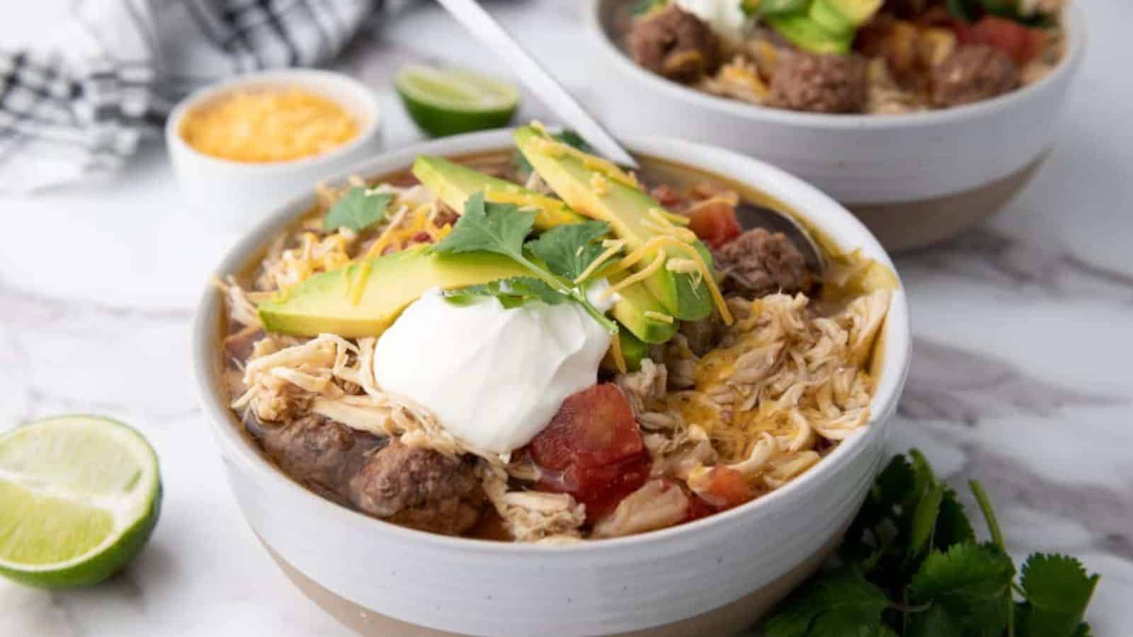 Two bowls of chili topped with avocado slices, sour cream, shredded cheese, and cilantro. Sliced lime and cheese bowl in the background on a marble surface.