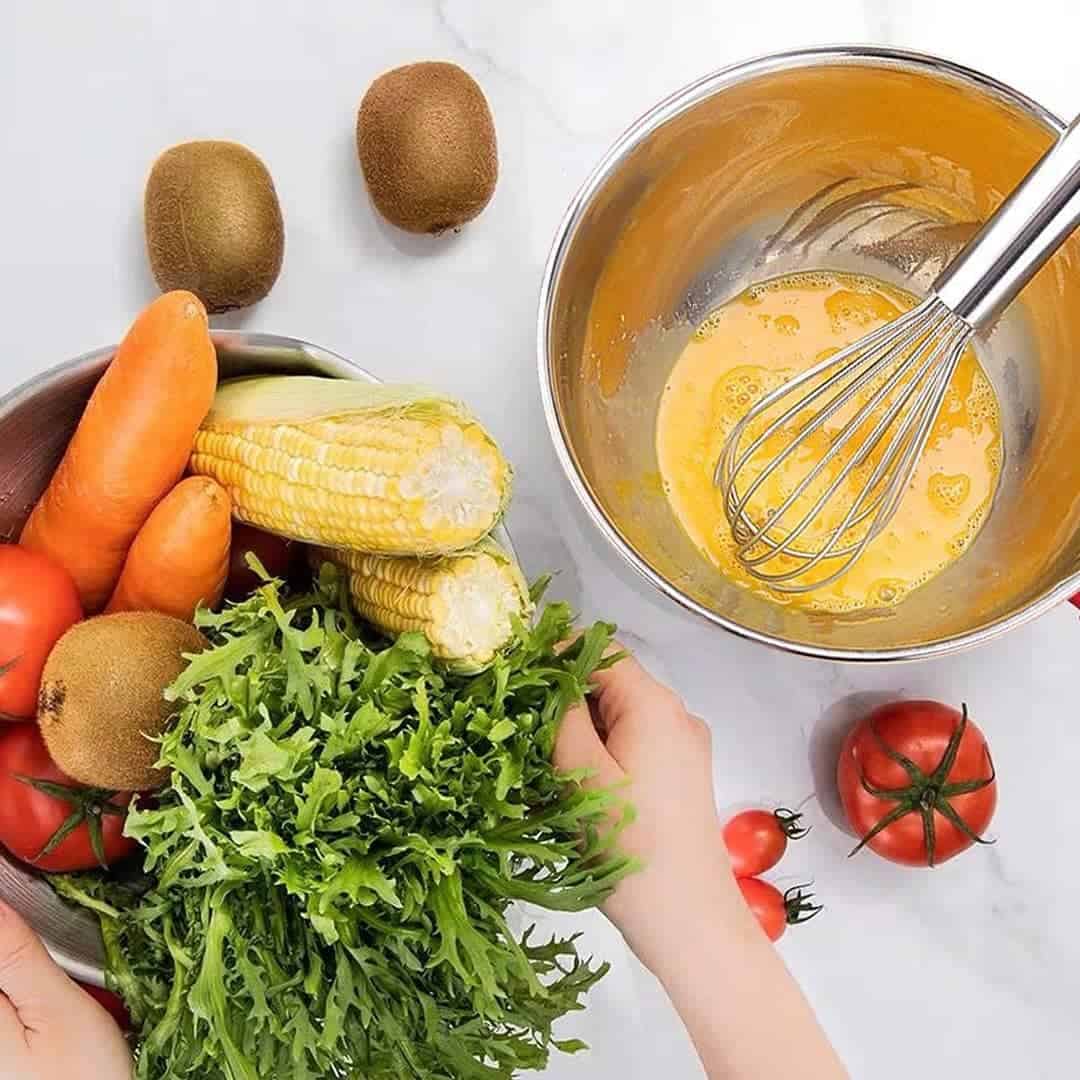Hands holding fresh vegetables next to a bowl of whisked eggs, corn, carrots, tomatoes, kiwis, and leafy greens.