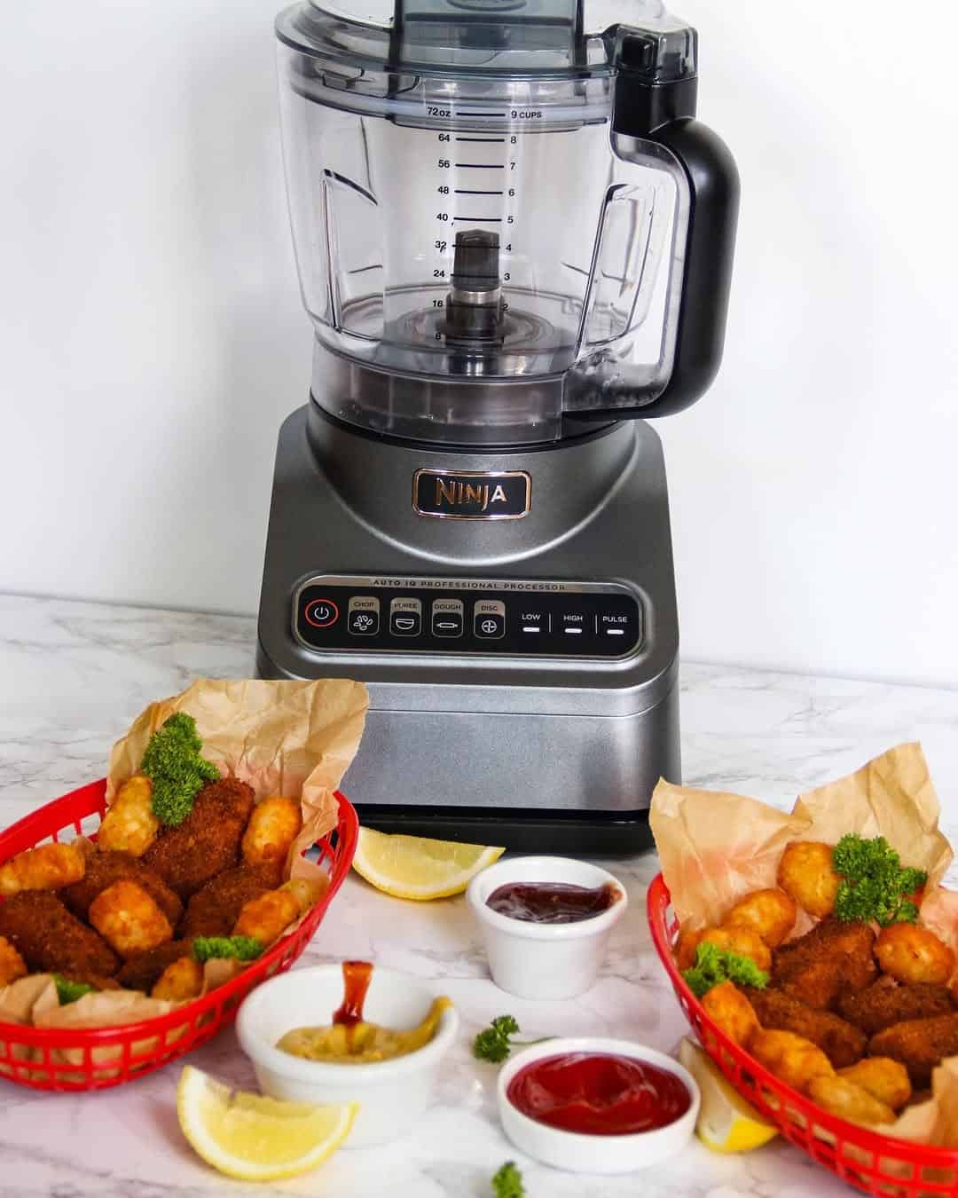 A Ninja blender stands behind baskets of fried snacks, sauces, and lemon wedges on a white countertop—one of the must-have kitchen gadgets for holiday cooking.
