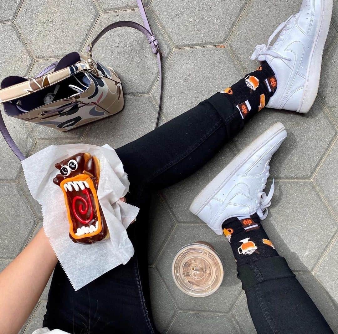 Woman sitting on pavement with a monster donut, iced drink, patterned socks, white sneakers, and a handbag—perfect inspiration for pumpkin spice gifts this season.