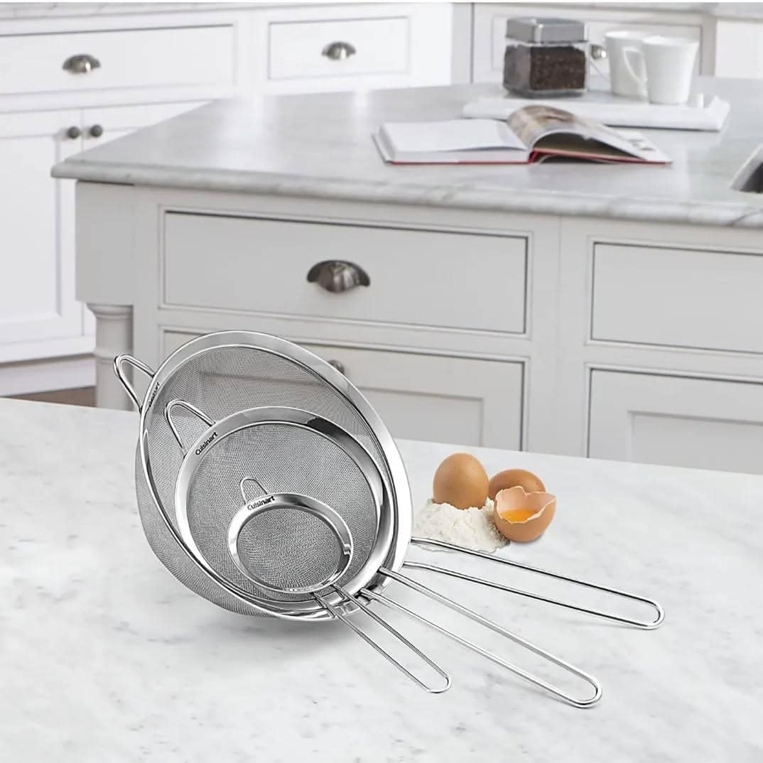Three metal mesh strainers, perfect kitchen tools for holiday baking, rest beside eggs and flour on a marble kitchen counter with white cabinets in the background.