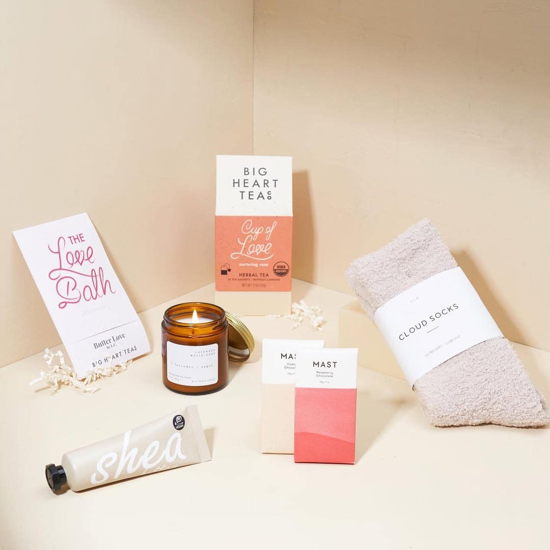 A cozy holiday gift set with tea, chocolate, a candle, lotion, socks, and a “Love Bath” card arranged on a beige surface—perfect for those seeking thoughtful and cozy holiday gifts.
