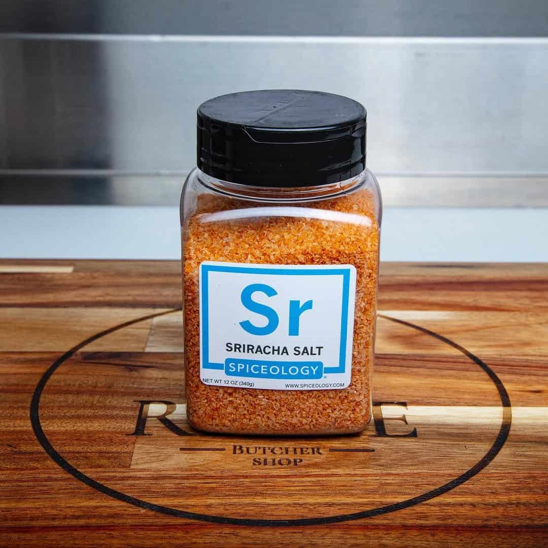 A jar of Spiceology Sriracha Salt sits on a wooden cutting board in a kitchen setting, making it an ideal addition to any Gifts for Holiday Hosts and Entertainers.
