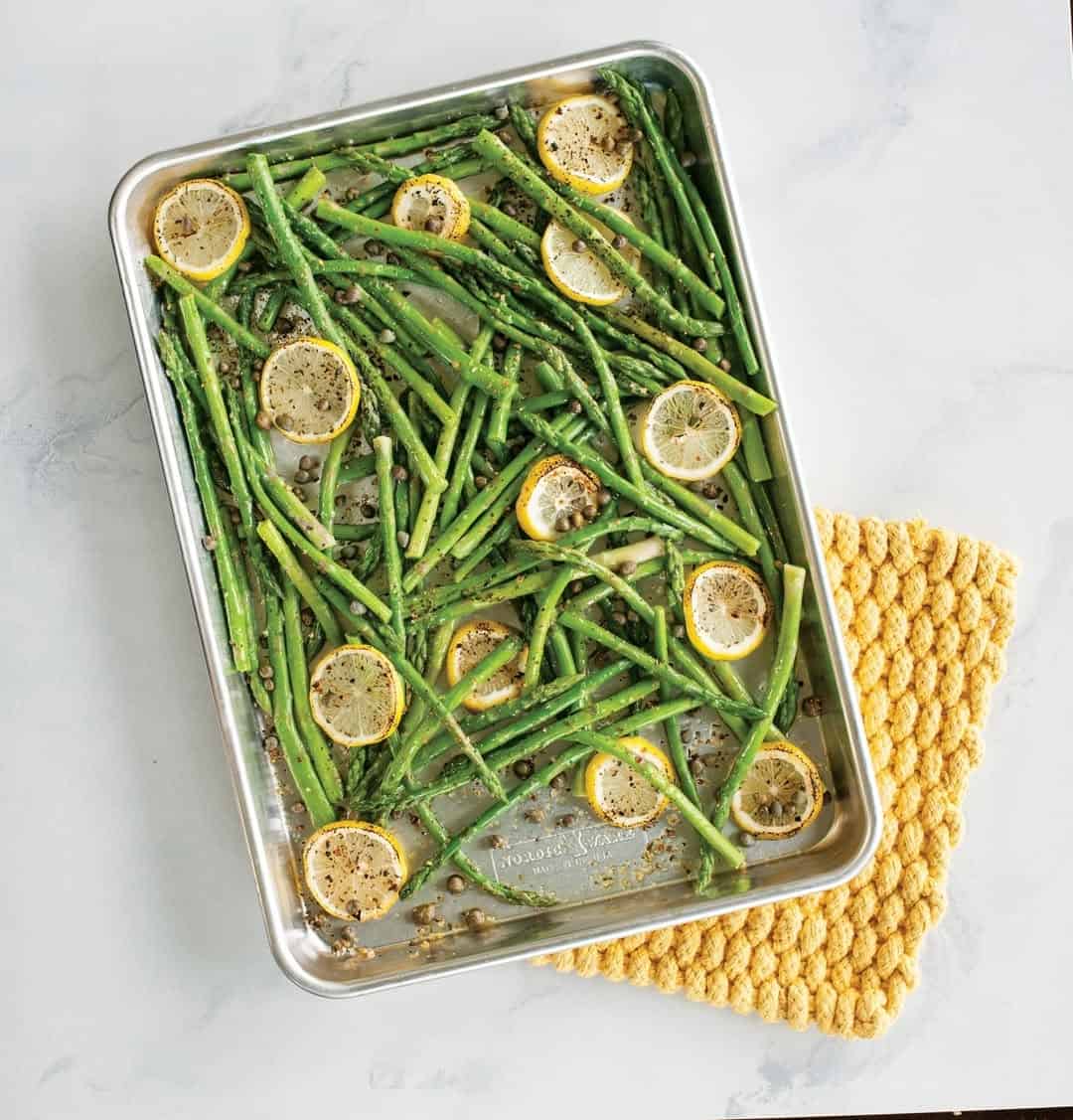 Roasted asparagus and lemon slices on a baking sheet, next to a yellow textured potholder&mdash;perfect inspiration for selecting kitchen tools for holiday baking.